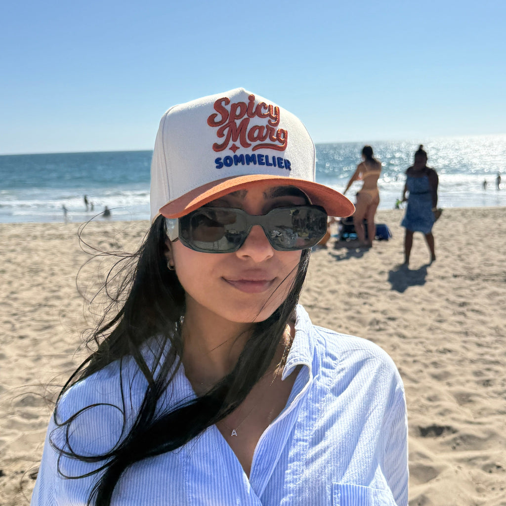 Woman wearing a 'Spicy Mals Sommelier' cap and sunglasses on a beach.