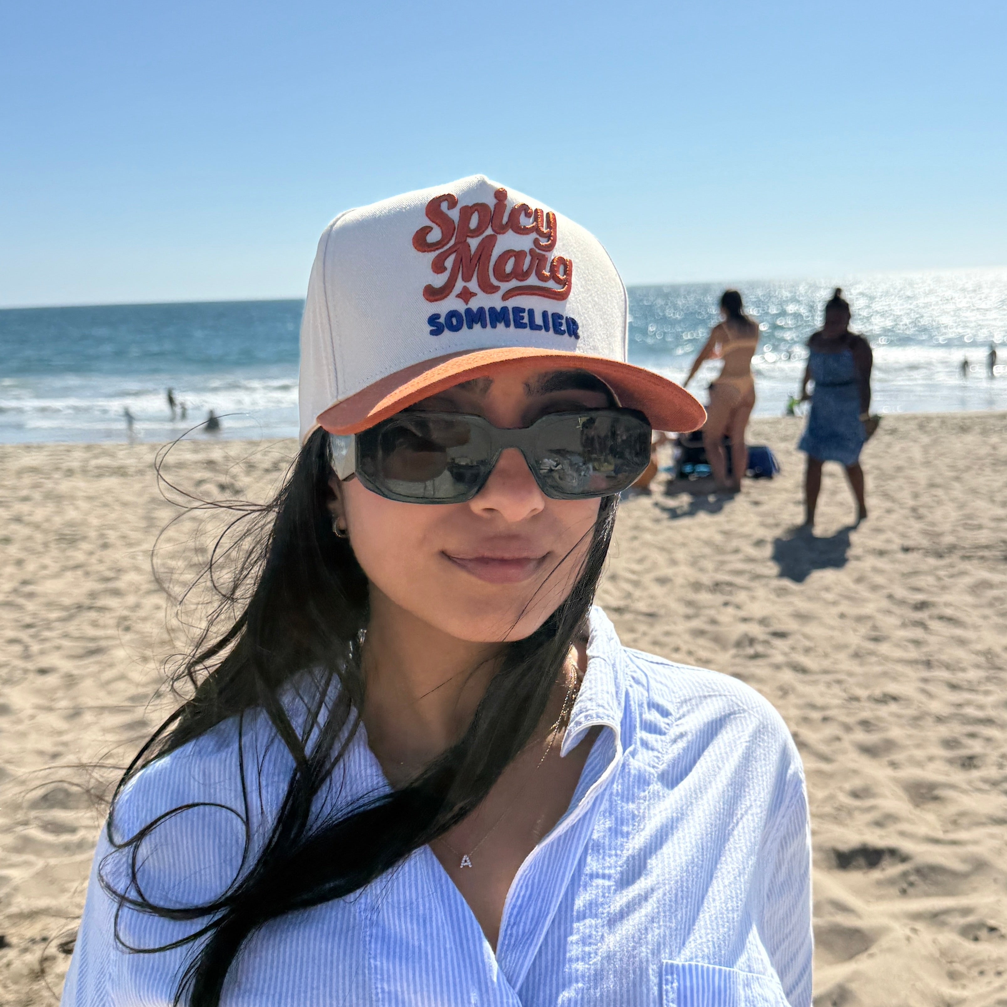 Woman wearing a 'Spicy Mals Sommelier' cap and sunglasses on a beach.