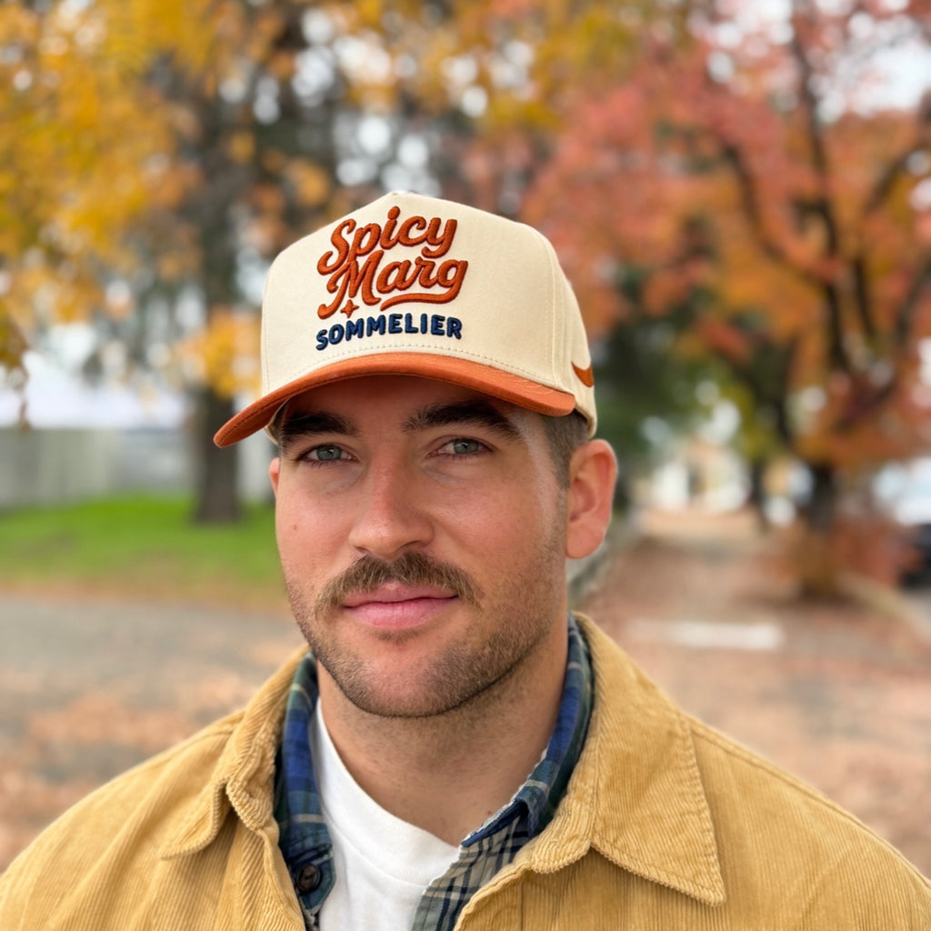 Man wearing a 'Spicy Marg Sommelier' cap with autumn trees in the background