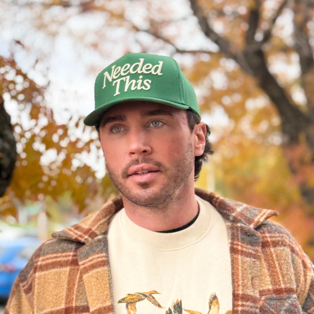 Man wearing a green cap and plaid jacket holding a drink, with autumn trees in the background